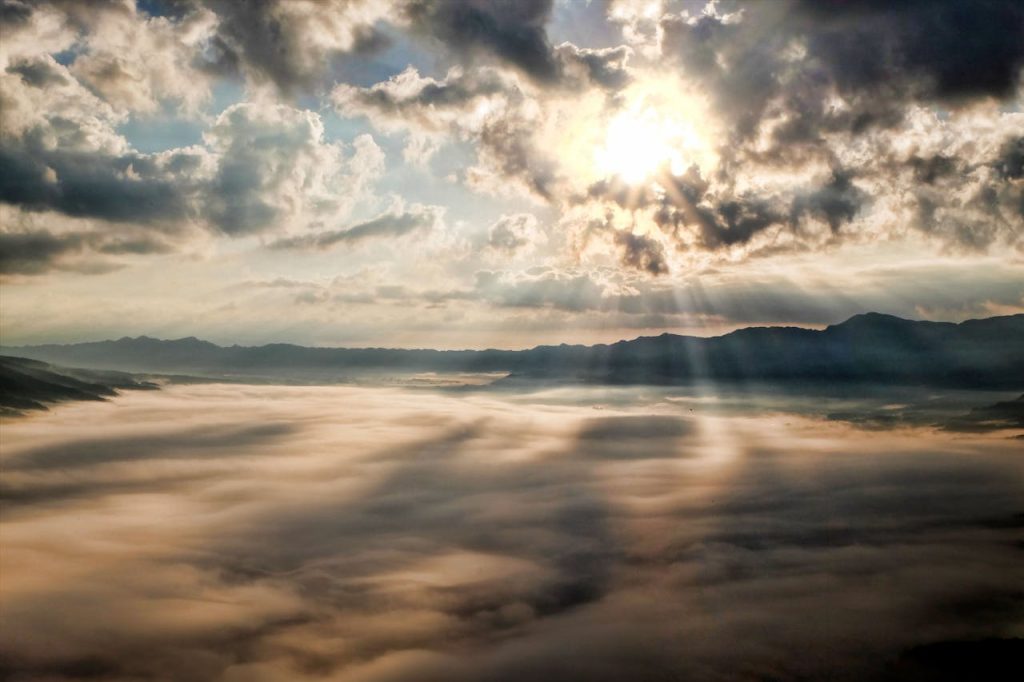 A landscape with clouds in the foreground and mountains in the background, the sun breaking through the clouds